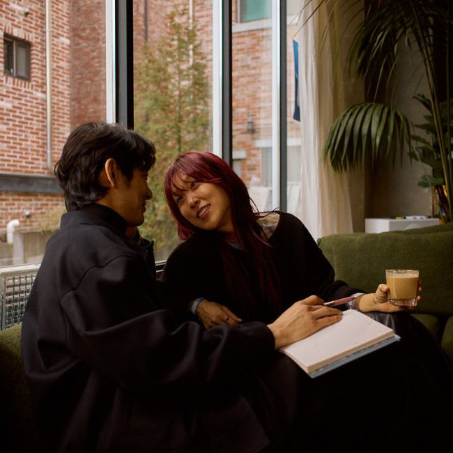 Two people in sitting by a window reading from an open book and drinking coffee